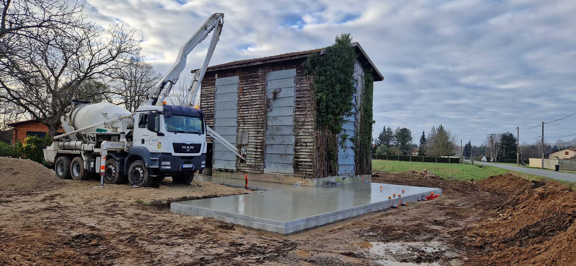 Camion malaxeur MAN verse du béton frais avec une pompe pour couler une dalle de fondation à côté d'une vieille grange en bois.