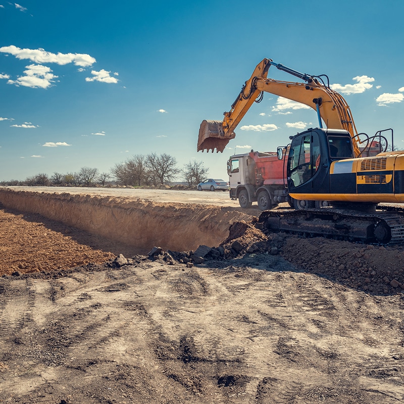 Excavatrice jaune creusant une longue tranchée de terre brune, chargeant une benne de camion sous un ciel bleu dégagé.