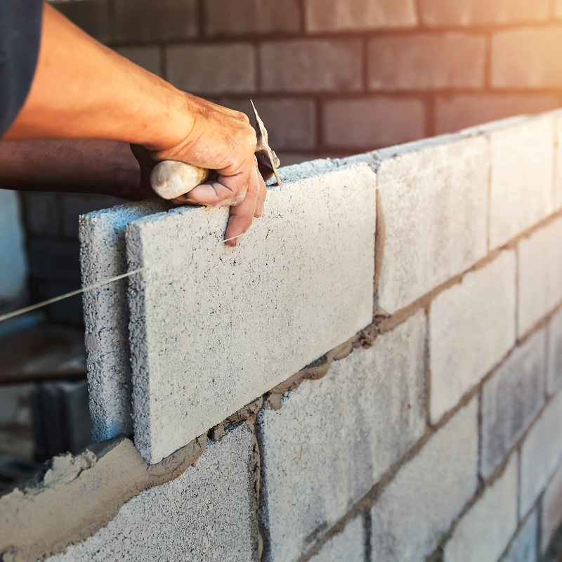 Mains d'un maçon posant un parpaing de béton cellulaire sur un lit de mortier, utilisant une truelle pour ajuster la construction du mur.