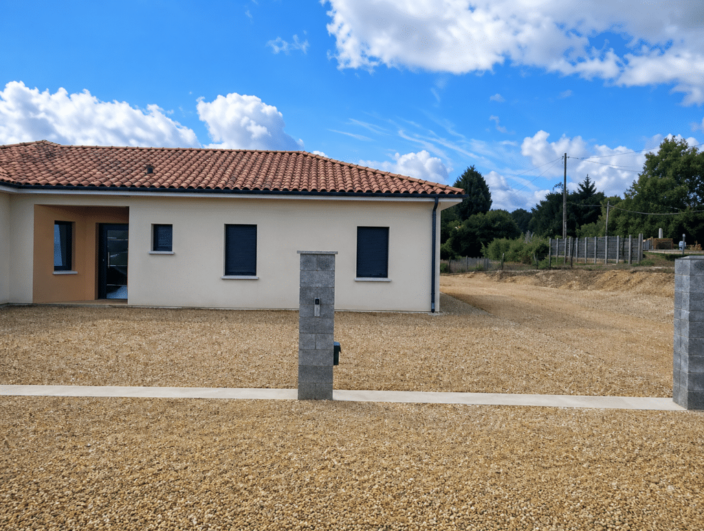 Maison moderne de plain-pied, façade beige, toit de tuiles rouges et cour gravillonnée sous un ciel bleu nuageux.