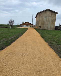 Long chemin de gravier jaune menant à une maison de campagne et un grand hangar en bois sous un ciel nuageux.
