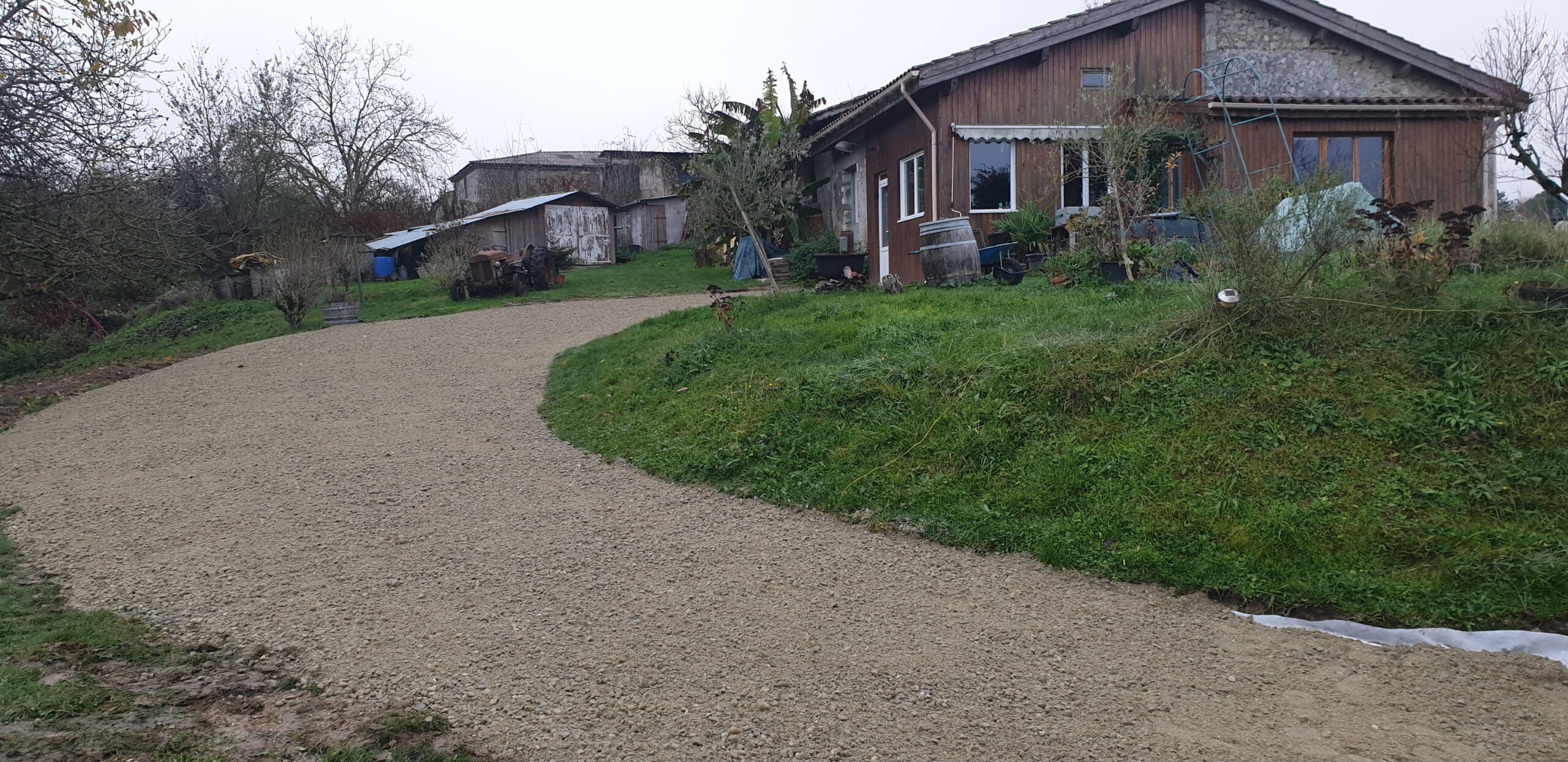 Allée en gravier fraîchement aménagée menant à une ferme rustique en bois avec un vieux tracteur en arrière-plan.