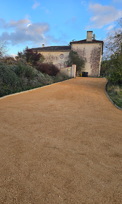 Allée en gravier ocre menant vers une maison de campagne ancienne en pierre sous un ciel bleu.