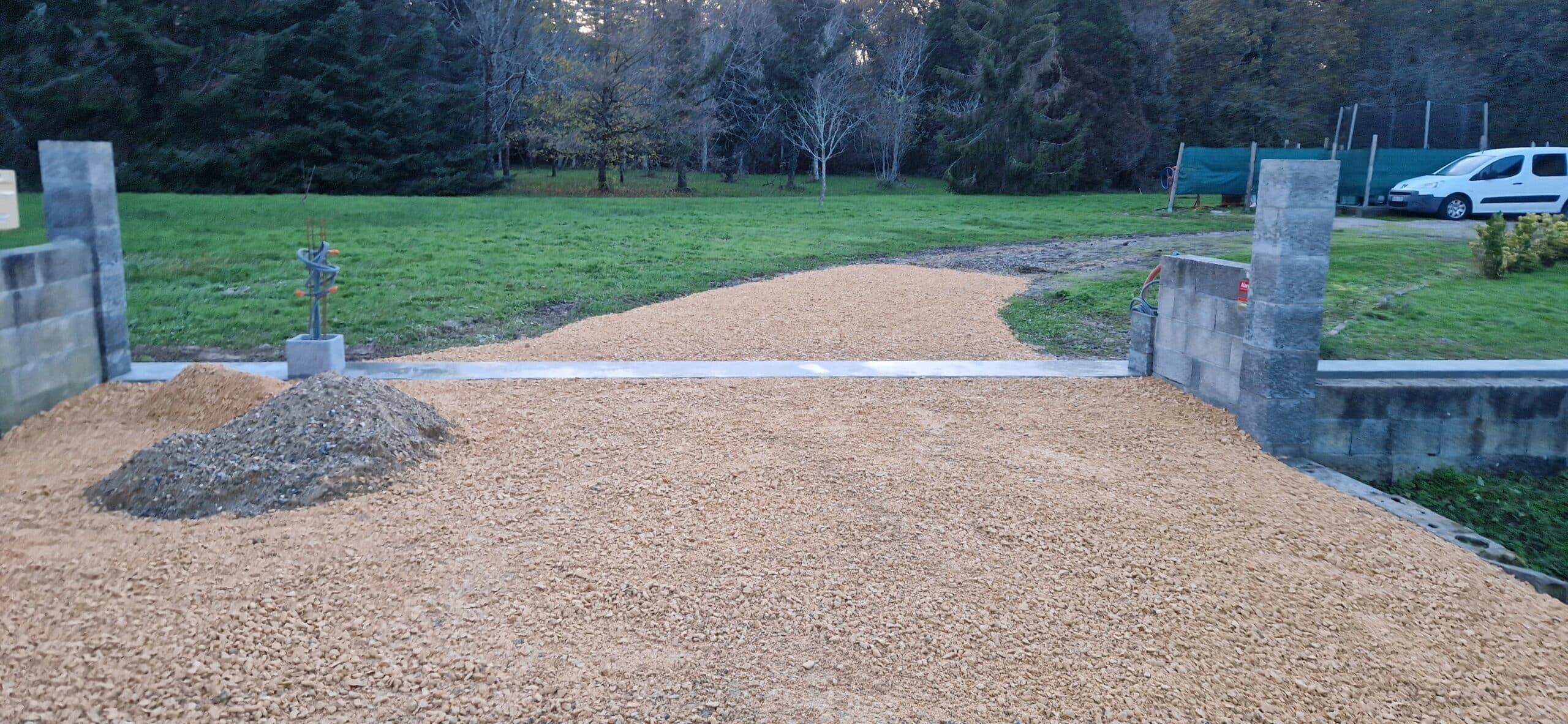 Chantier d'allée avec gravier beige, seuil en béton et piliers en parpaings devant une forêt.