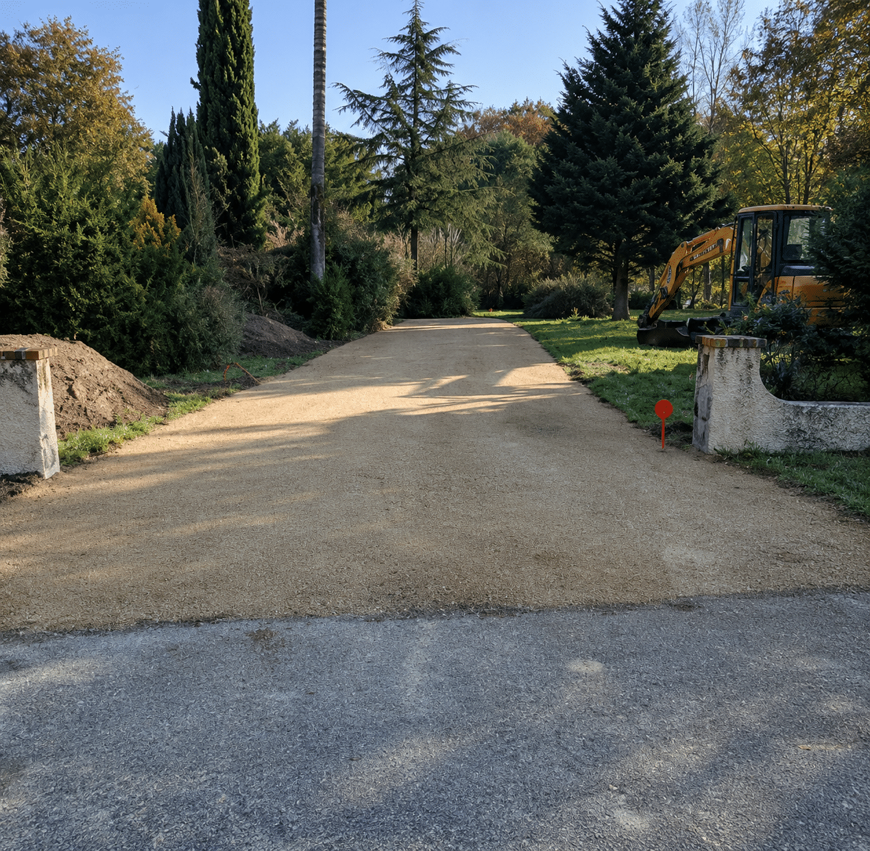 Aménagement d’une Allée en Gravier pour Jardin Paysager Aménagement d'une allée en gravier beige menant à un parc boisé avec une pelleteuse jaune en bordure.