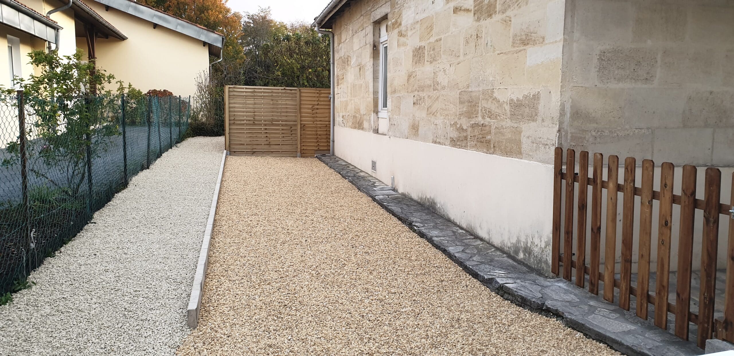 Aménagement extérieur avec allée de gravier ocre, bordure en pierre naturelle et clôture bois devant une maison ancienne.