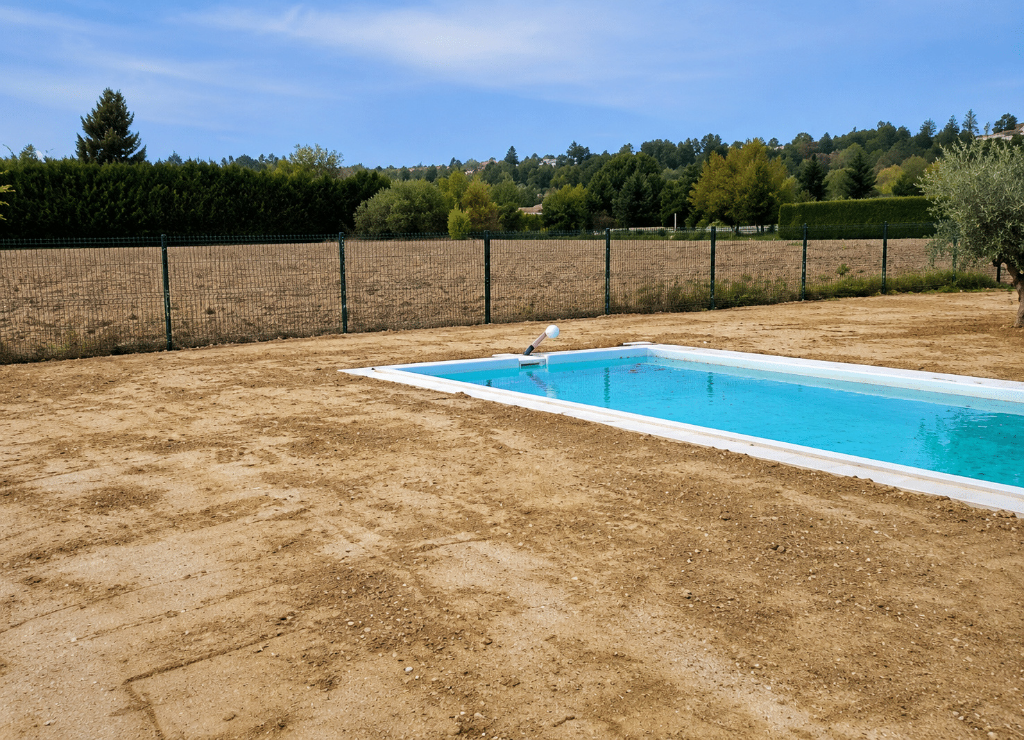 Piscine bleue installée sur un terrain en terre avec clôture grillagée et paysage verdoyant en arrière-plan.