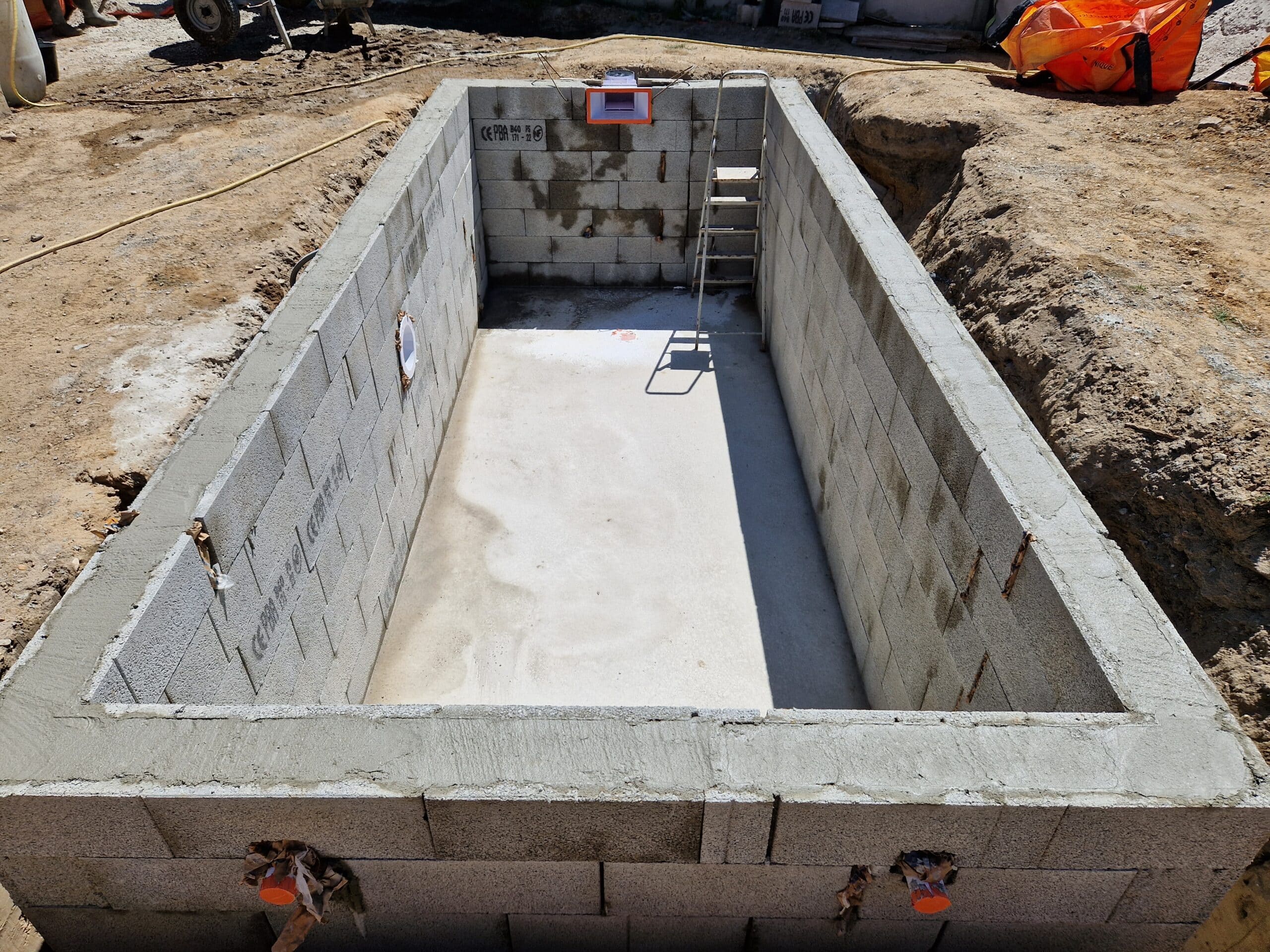 Construction d'une piscine rectangulaire en blocs béton avec skimmer et dalle apparente sur un chantier de terrassement.