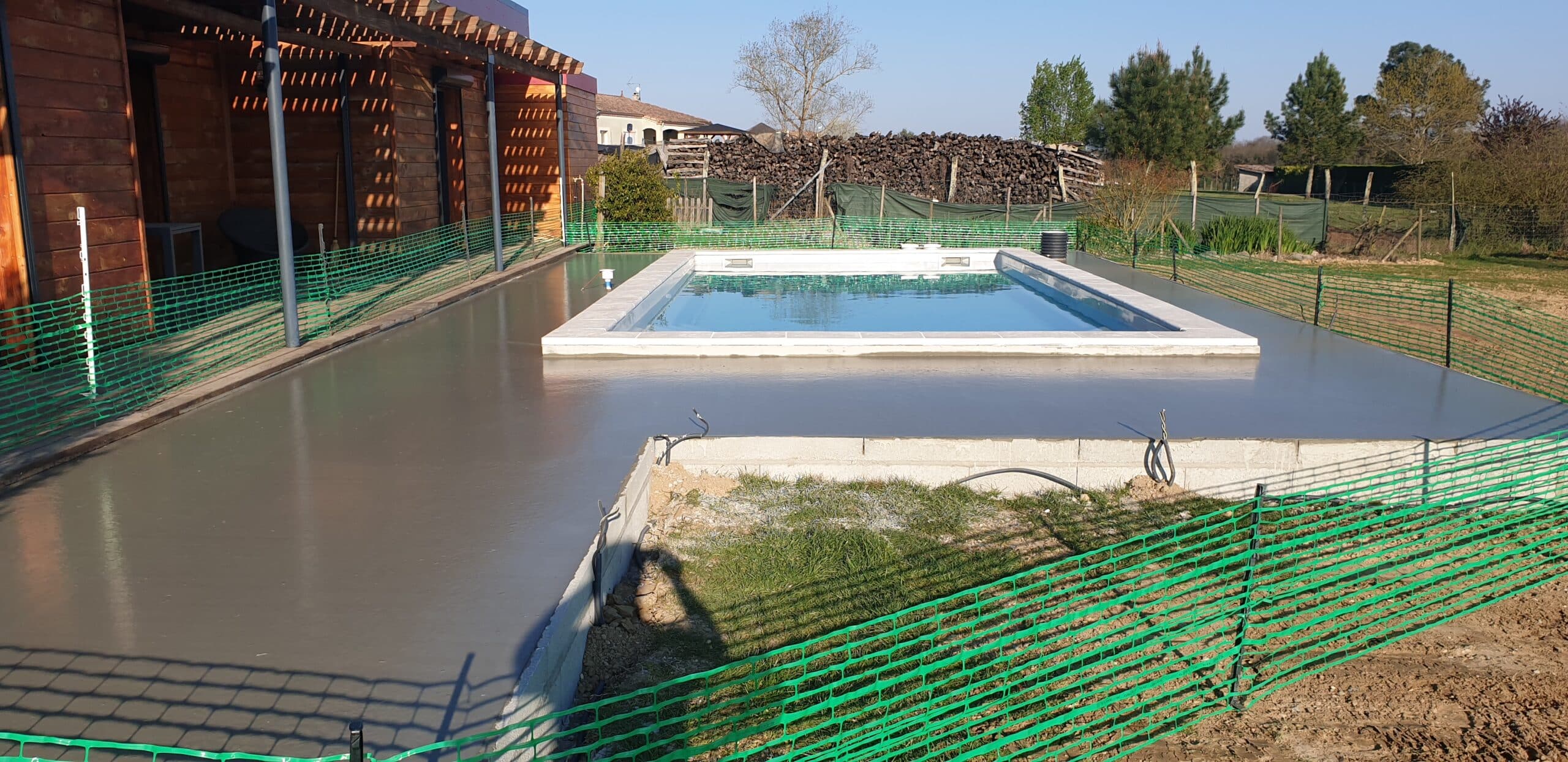 Terrasse en béton frais autour d'une piscine enterrée en cours de construction avec clôture de sécurité verte.