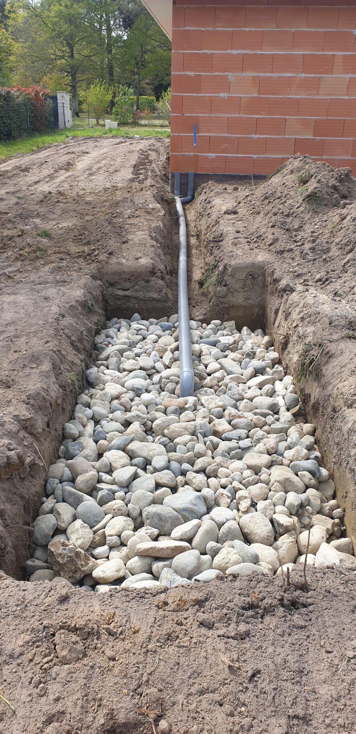 Installation d'un puits perdu avec tuyau PVC et lit de galets pour le drainage des eaux pluviales d'une maison.