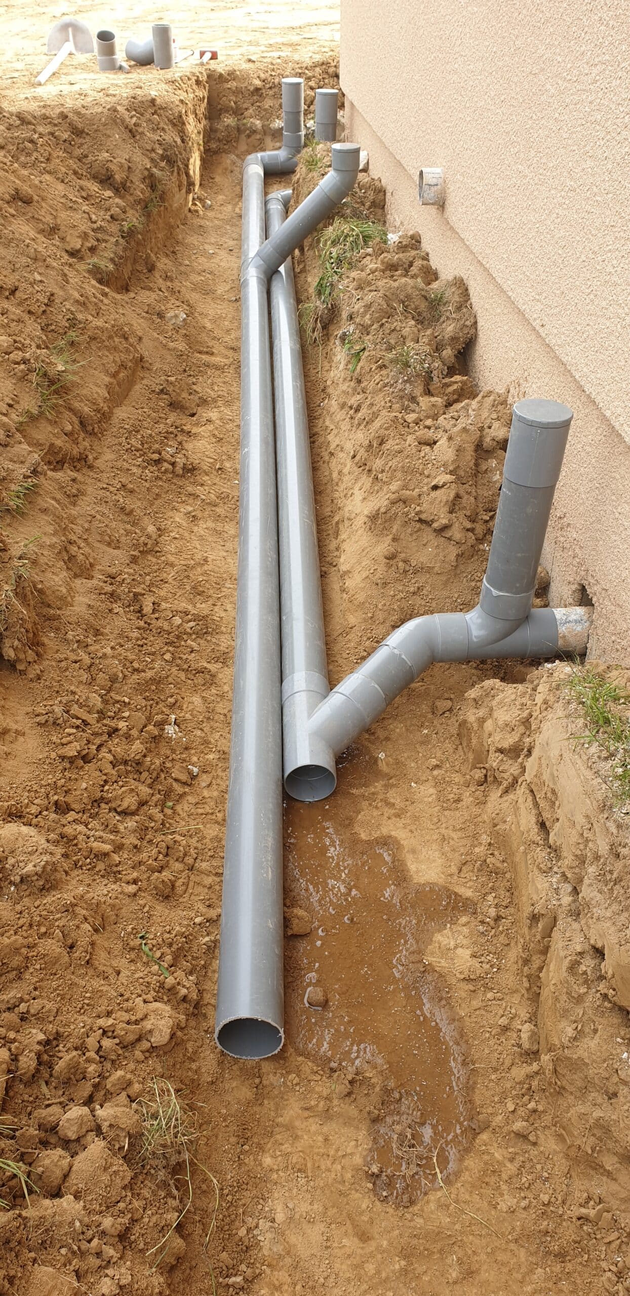 Installation de tuyaux de drainage en PVC gris dans une tranchée devant le mur extérieur d'une maison en chantier.
