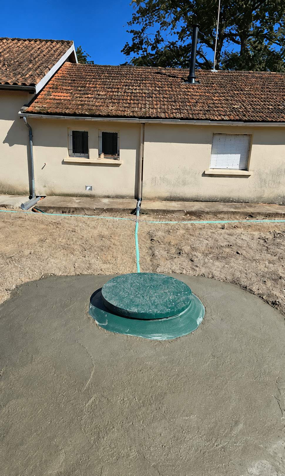 Installation de fosse septique : couvercle vert et béton Couvercle de fosse septique vert sur béton frais devant une maison de plain-pied avec toiture en tuiles.