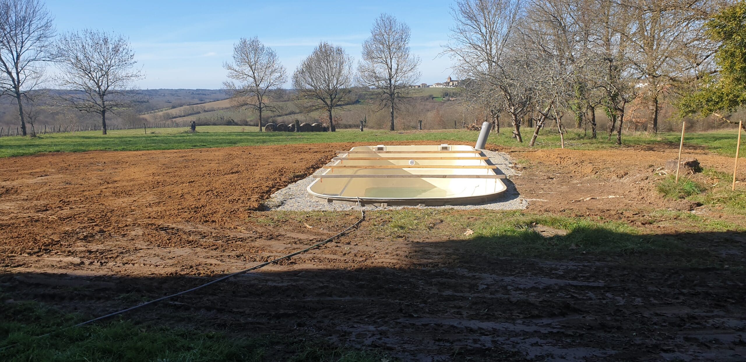 Coque de piscine beige en cours de pose avec renforts bois sur lit de gravier dans un paysage rural.