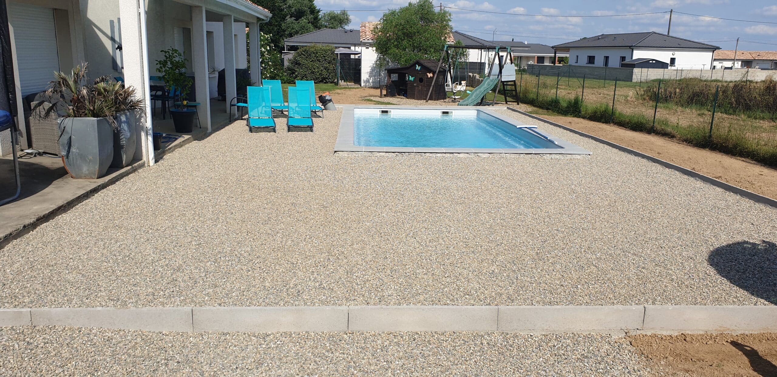 Piscine rectangulaire entourée d'une vaste terrasse en gravier avec transats bleus et aire de jeux au fond du jardin.
