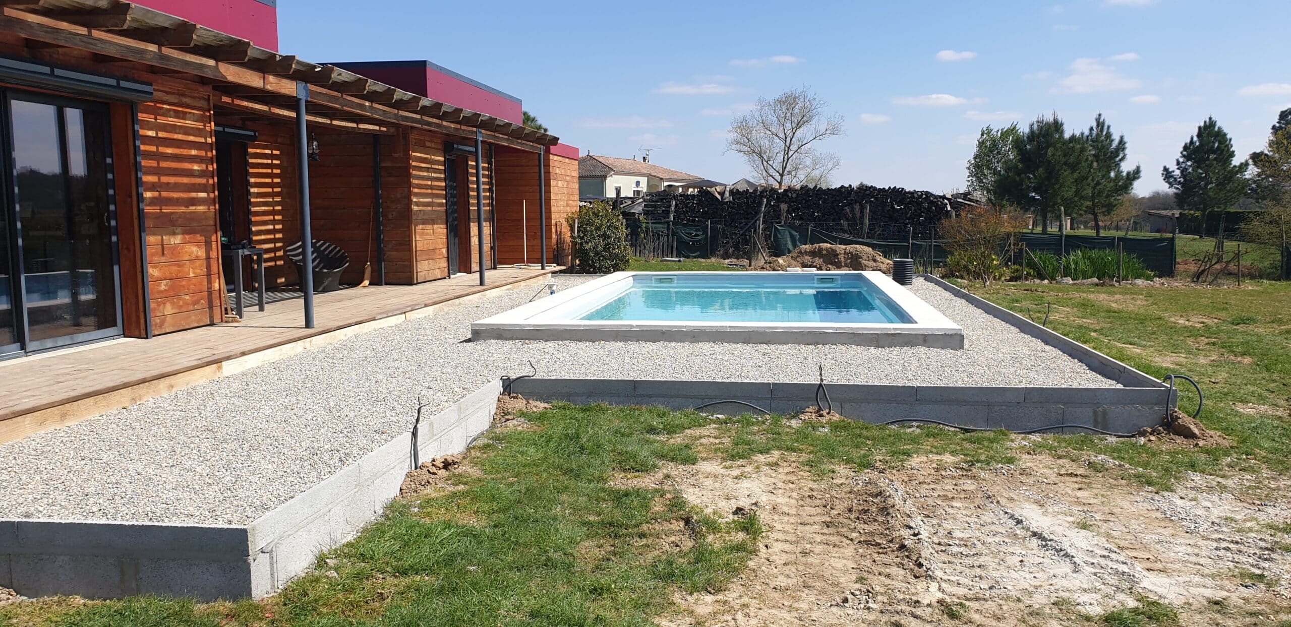 Piscine moderne avec terrasse en bois et plage de gravier blanc devant une maison contemporaine en bois.