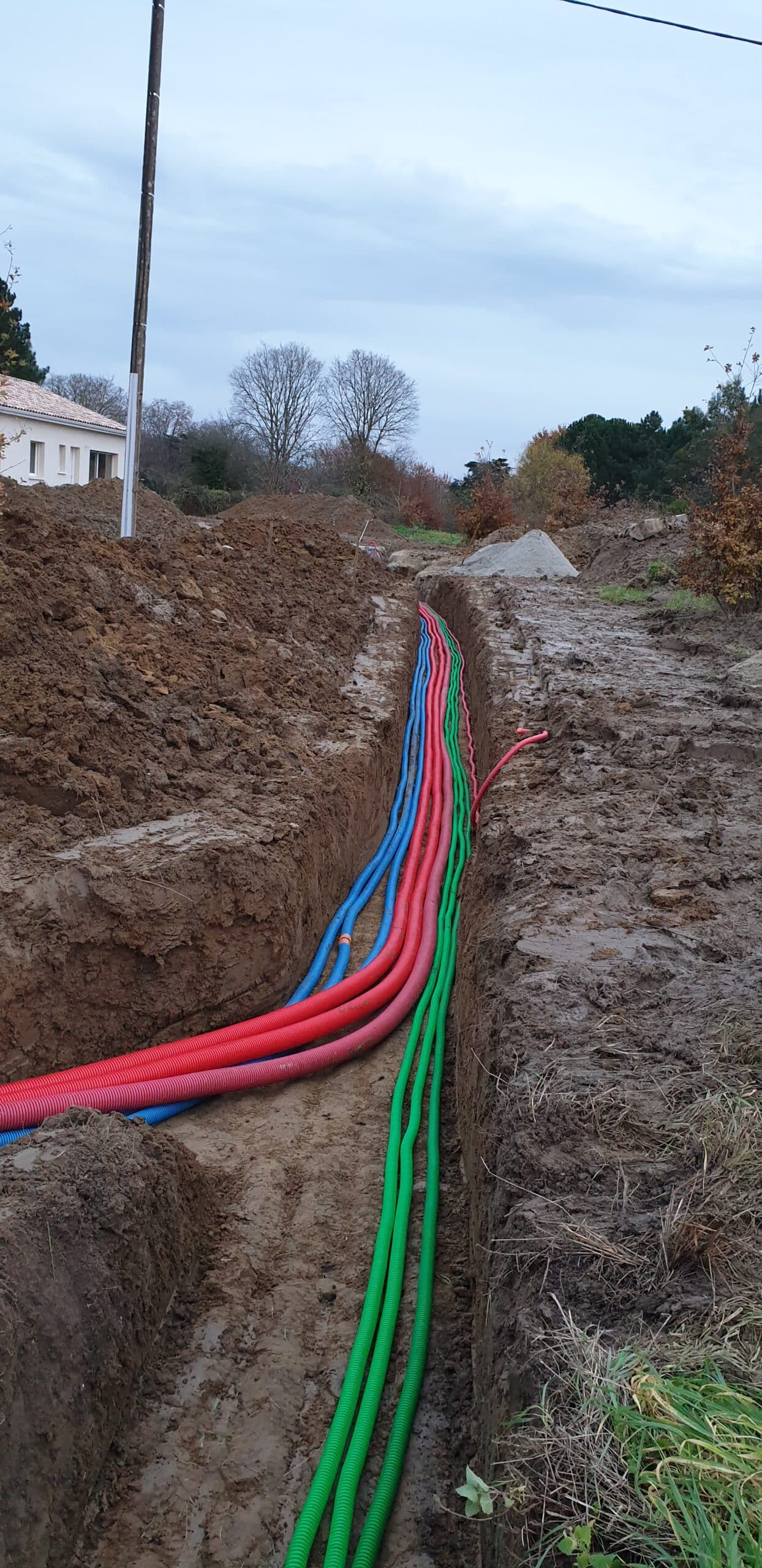 Réseaux de gaines électriques et télécom colorées posés dans une tranchée de chantier boueuse.
