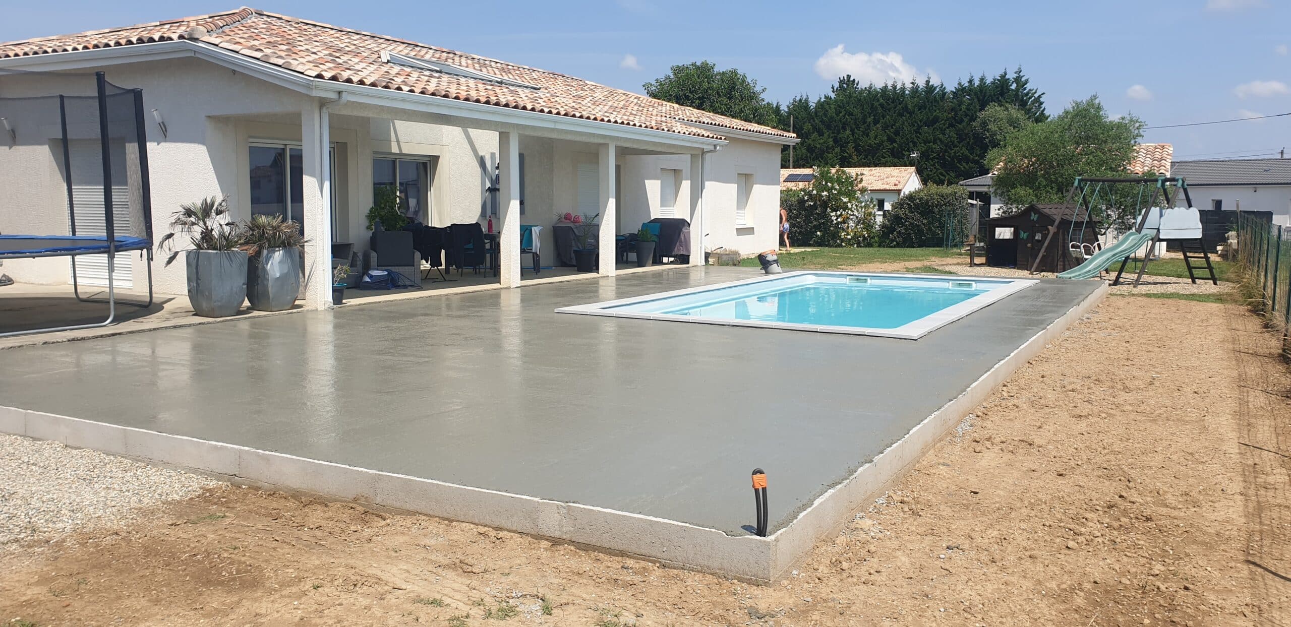 Terrasse en béton fraîchement coulée autour d'une piscine devant une maison moderne avec jardin et pergola.
