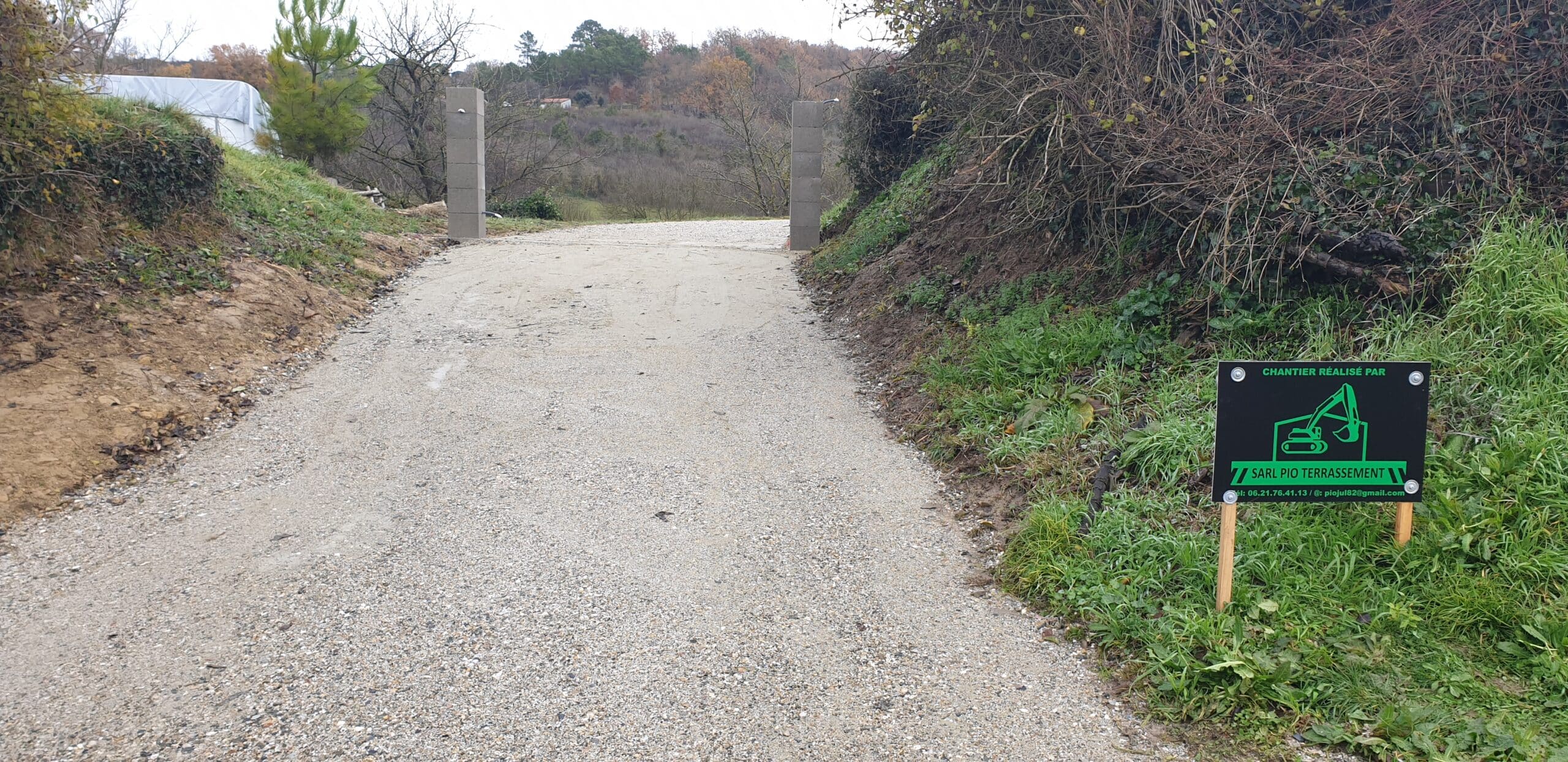 Chemin gravillonné avec panneau SARL PIO Terrassement devant deux piliers d'entrée en béton gris.