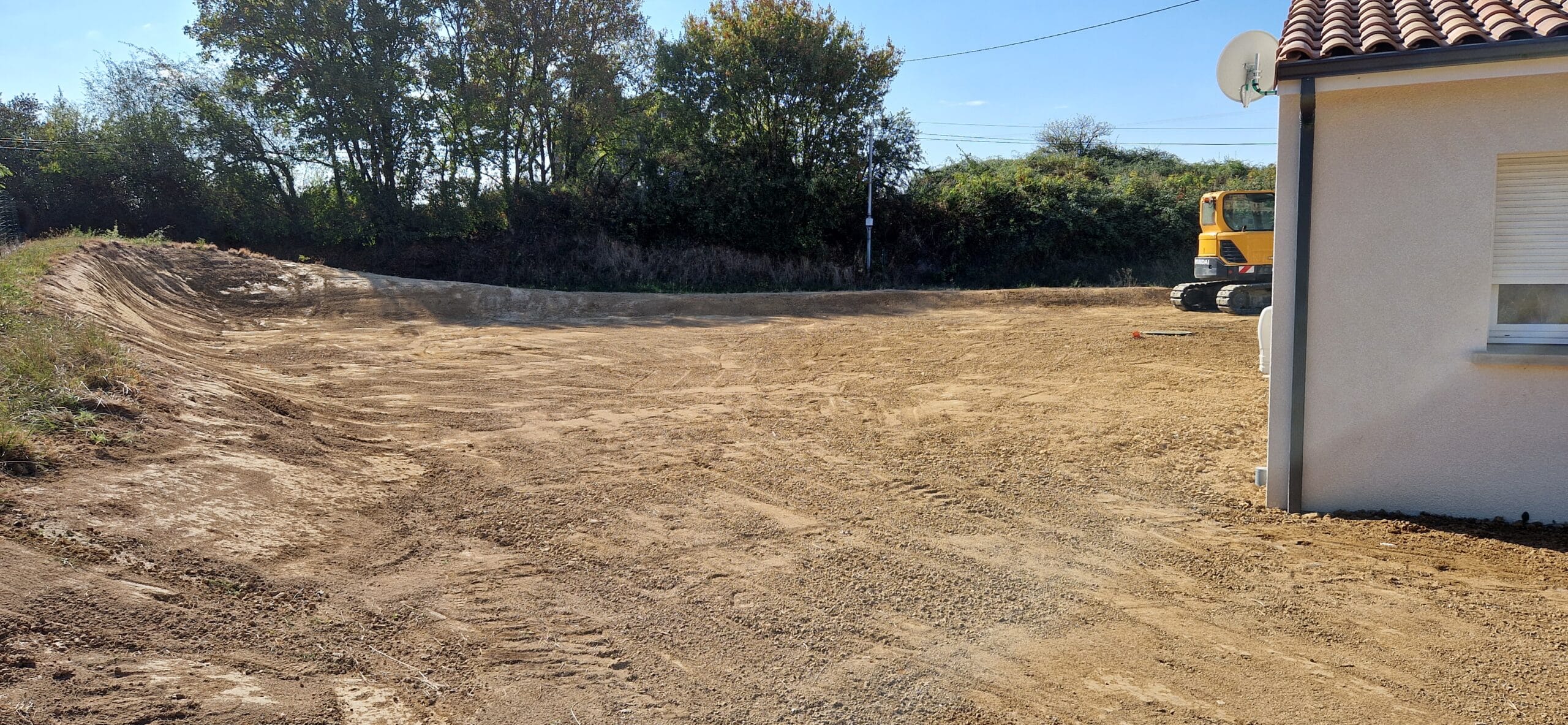 Terrain en cours de terrassement à côté d'une maison avec une pelleteuse Hyundai en arrière-plan.
